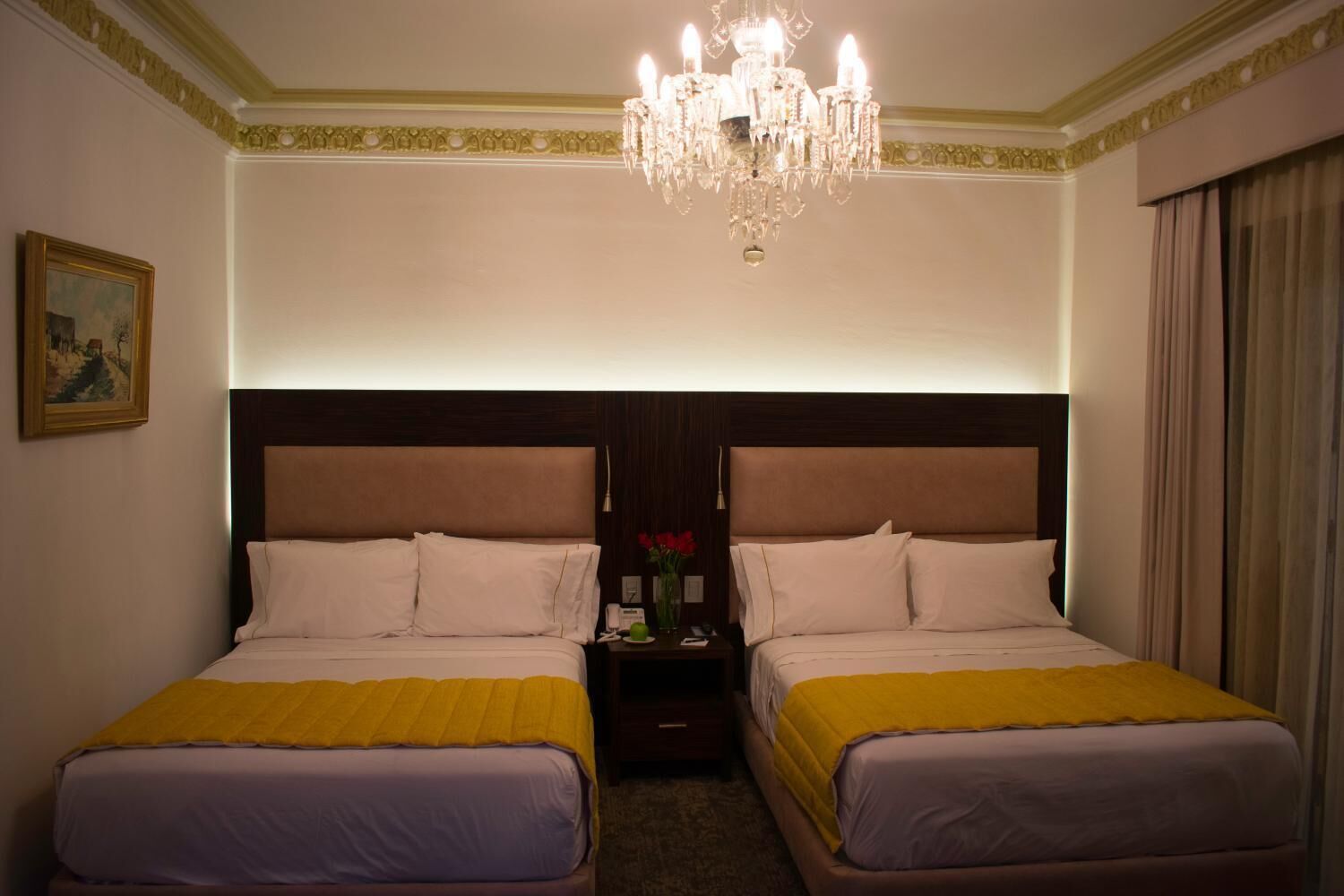 Een hotelkamer met twee bedden, een kroonluchter, een schilderij en een nachtkastje met een bloemenvaas.