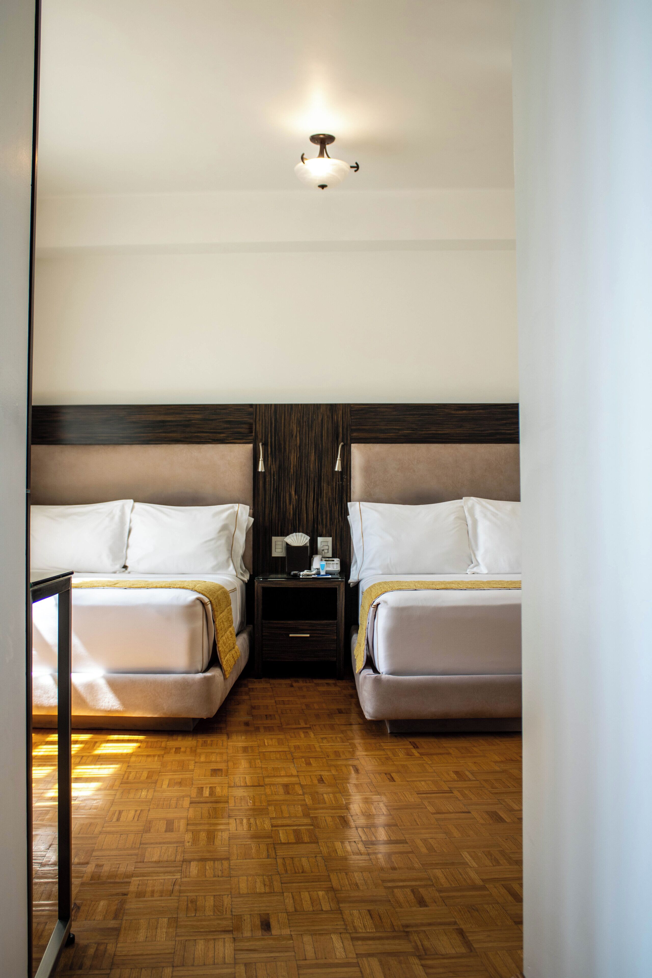 A hotel room with two beds, a wooden floor, a nightstand, and a ceiling light.