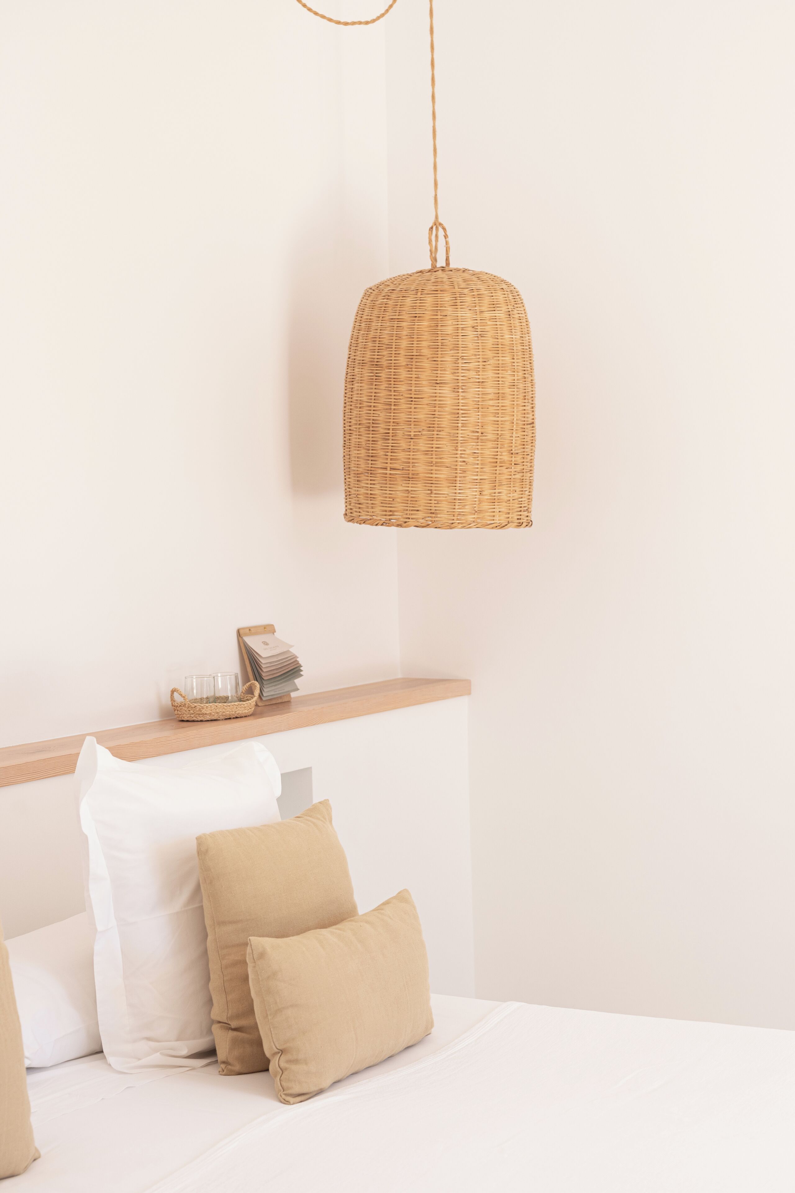 Une chambre avec un lit blanc, des oreillers beige, une table de chevet en bois, une suspension en osier et un mur blanc.