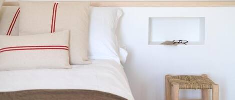 A neatly made bed with white bedding and red-striped pillows, a small wooden stool, and a framed picture on the wall.