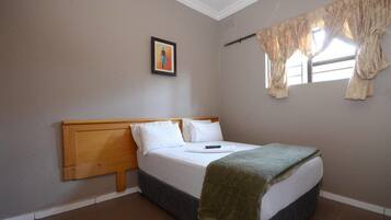 Une chambre à coucher avec une tête de lit en bois, un lit recouvert de draps blancs, un plaid vert, un tableau encadré accroché au mur et une fenêtre avec des rideaux.