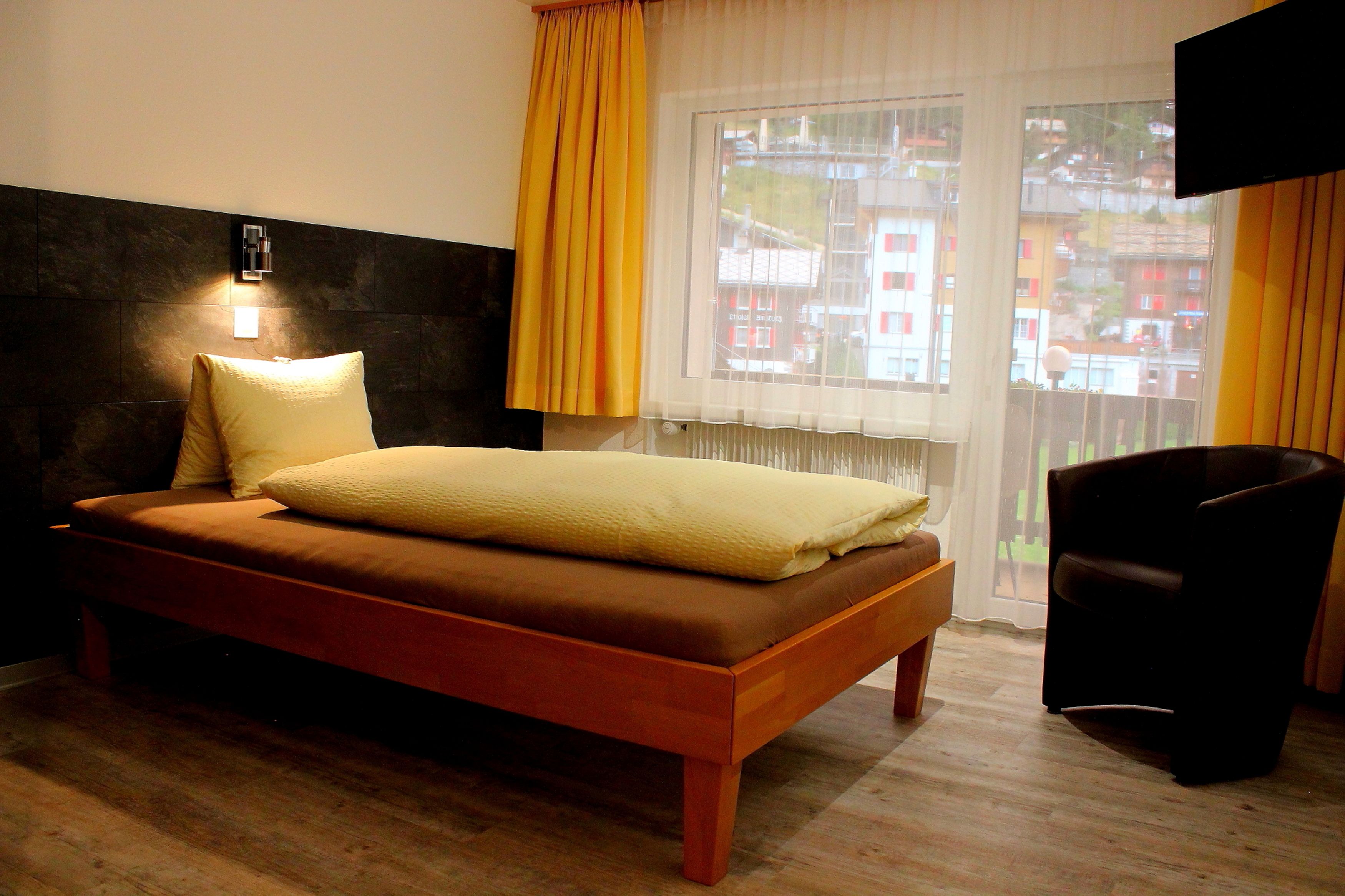 A bedroom with a bed, a bedside table, a chair, a window with curtains, and a wall-mounted TV.