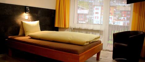 A bedroom with a bed, a bedside table, a chair, a window with curtains, and a wall-mounted TV.