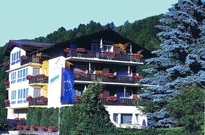 A multi-story hotel with balconies adorned with red flowers, a visible sign reading "Garten Alpha," and a lush green backdrop.