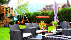 A patio area with wicker furniture, a small table with a potted plant, and a swing set in the background. - Alpha (Graechen)