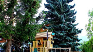 A wooden play structure with a slide and a swing set in a grassy area with trees. - Alpha (Graechen)