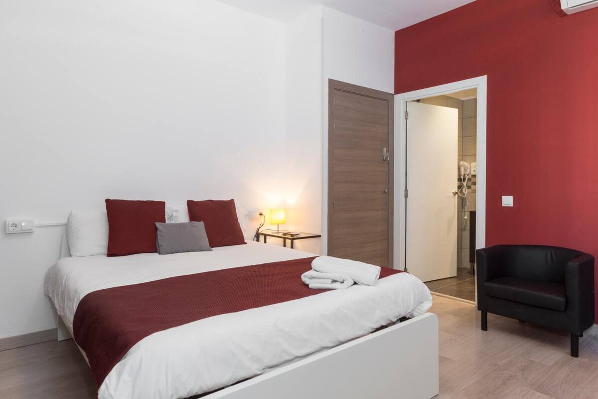A modern hotel room with a large bed, red and white bedding, a side table with a lamp, and a black armchair.