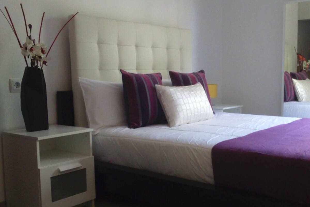 A bedroom with a white bed, purple and white pillows, a white nightstand, and a purple throw blanket.