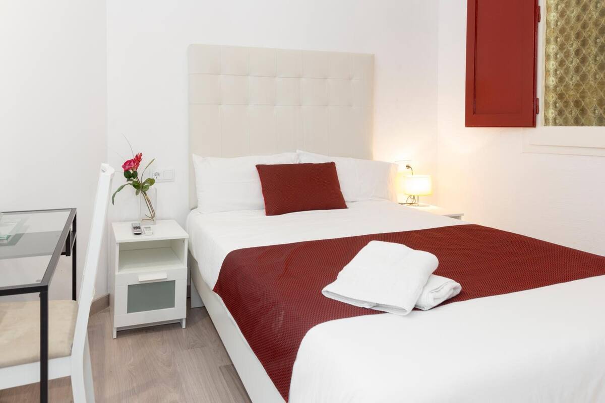A hotel room with a bed, bedside table, lamp, and a small table with a vase of flowers.