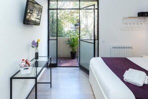 A hotel room with a bed, a small table with a vase of flowers, a potted plant, and a sliding door leading to an outdoor area with a view of trees and a balcony. - Brustar Sagrada Familia (Barcelona)