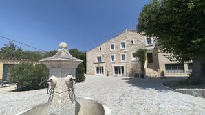 A stone fountain with a spherical top and two spouts, surrounded by cobblestone pavement, in front of a stone building with multiple windows and a door.