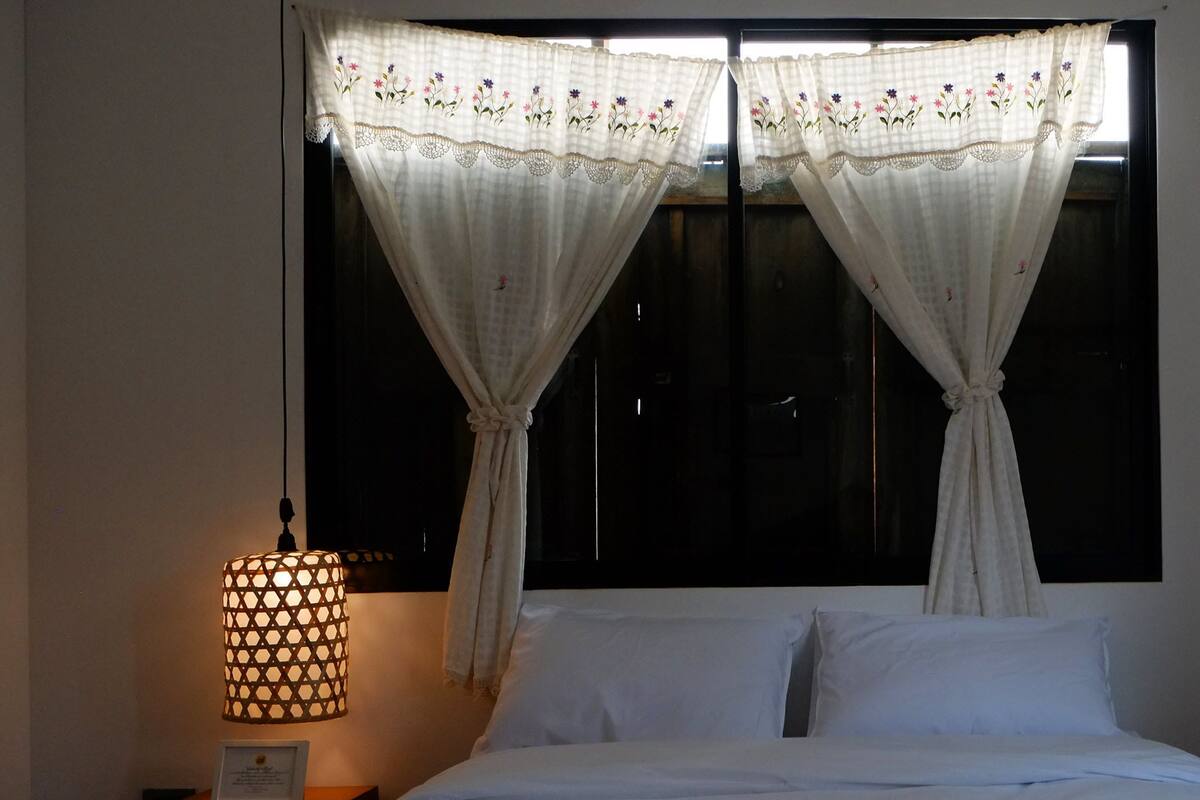 A bedroom with a bed, white curtains, a bedside lamp, and a window.