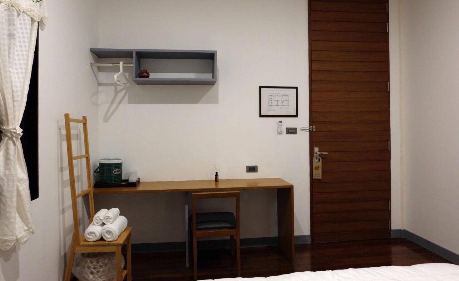 A bedroom with a wooden desk, a chair, a bed, a wooden wardrobe, and a small shelf with a vase.