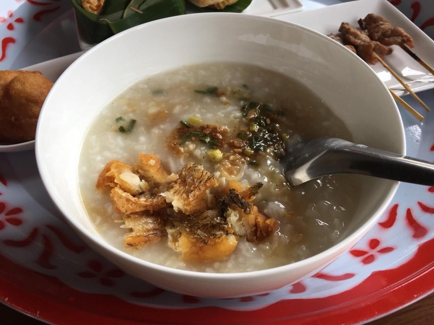 Un tazón de sopa con acompañamientos fritos y una cuchara, servido en un plato blanco con un diseño rojo.