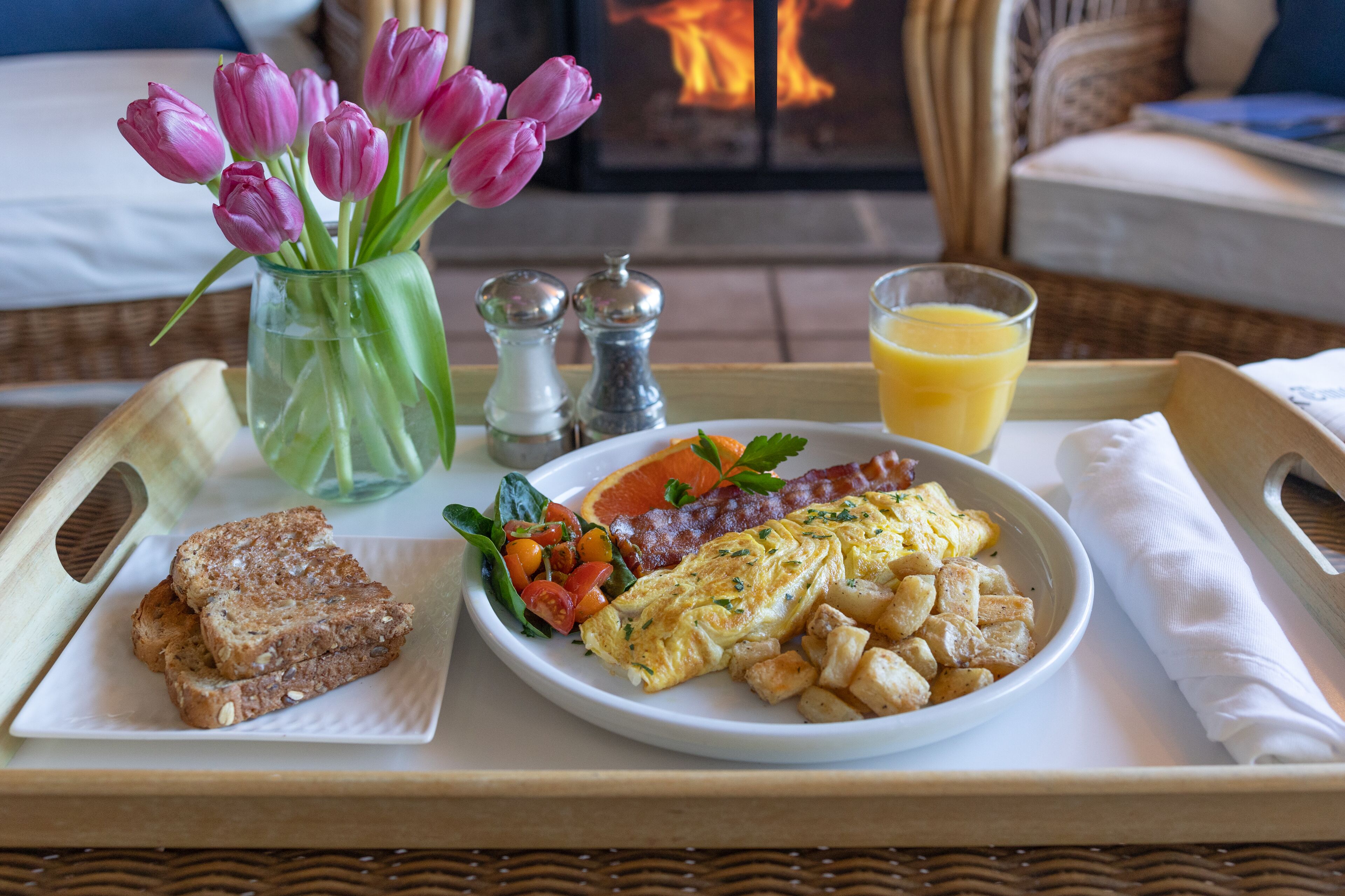 A tray with breakfast items including toast, eggs, potatoes, tomatoes, and a glass of orange juice, with a vase of pink tulips and salt and pepper shakers in the background.