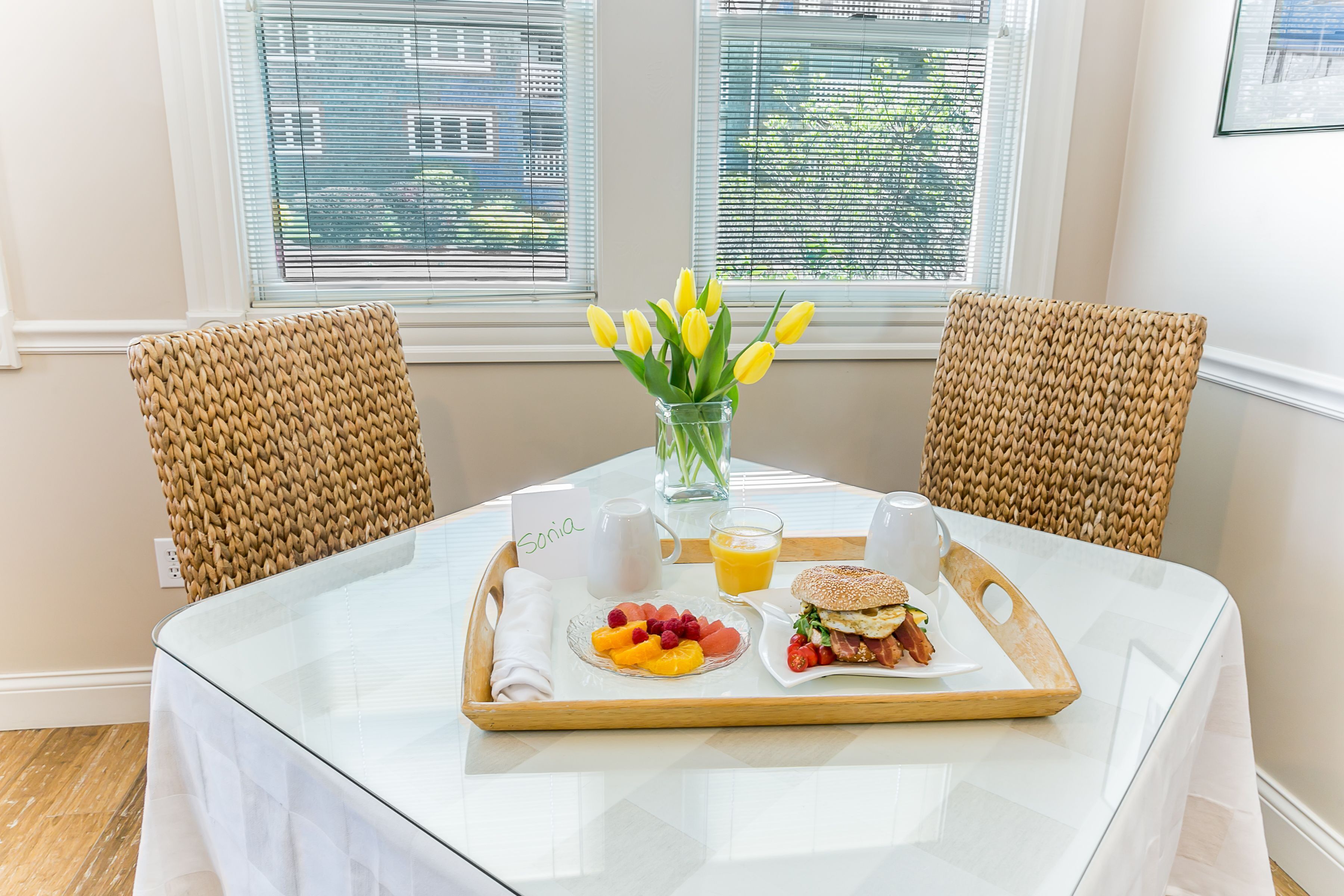 A breakfast tray on a table with a glass of orange juice, a plate of fruit, and a sandwich, with a vase of yellow flowers in the center.