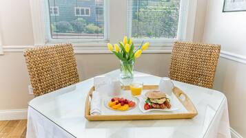 A breakfast tray on a table with a glass of orange juice, a plate of fruit, and a sandwich, with a vase of yellow flowers in the center.
