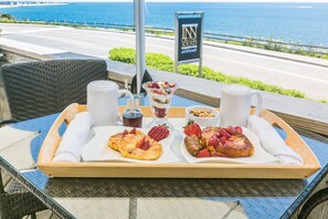 A breakfast tray with pancakes, strawberries, and a dessert on a table with a view of the ocean.
