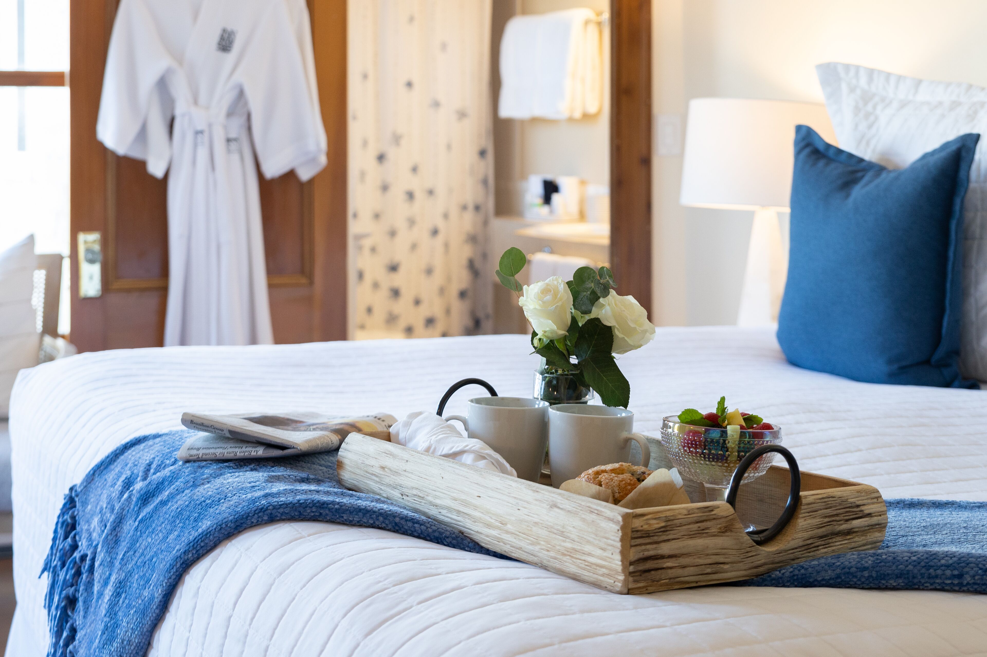 A neatly made bed with a tray of breakfast items, a vase of flowers, and a robe hanging on the door.
