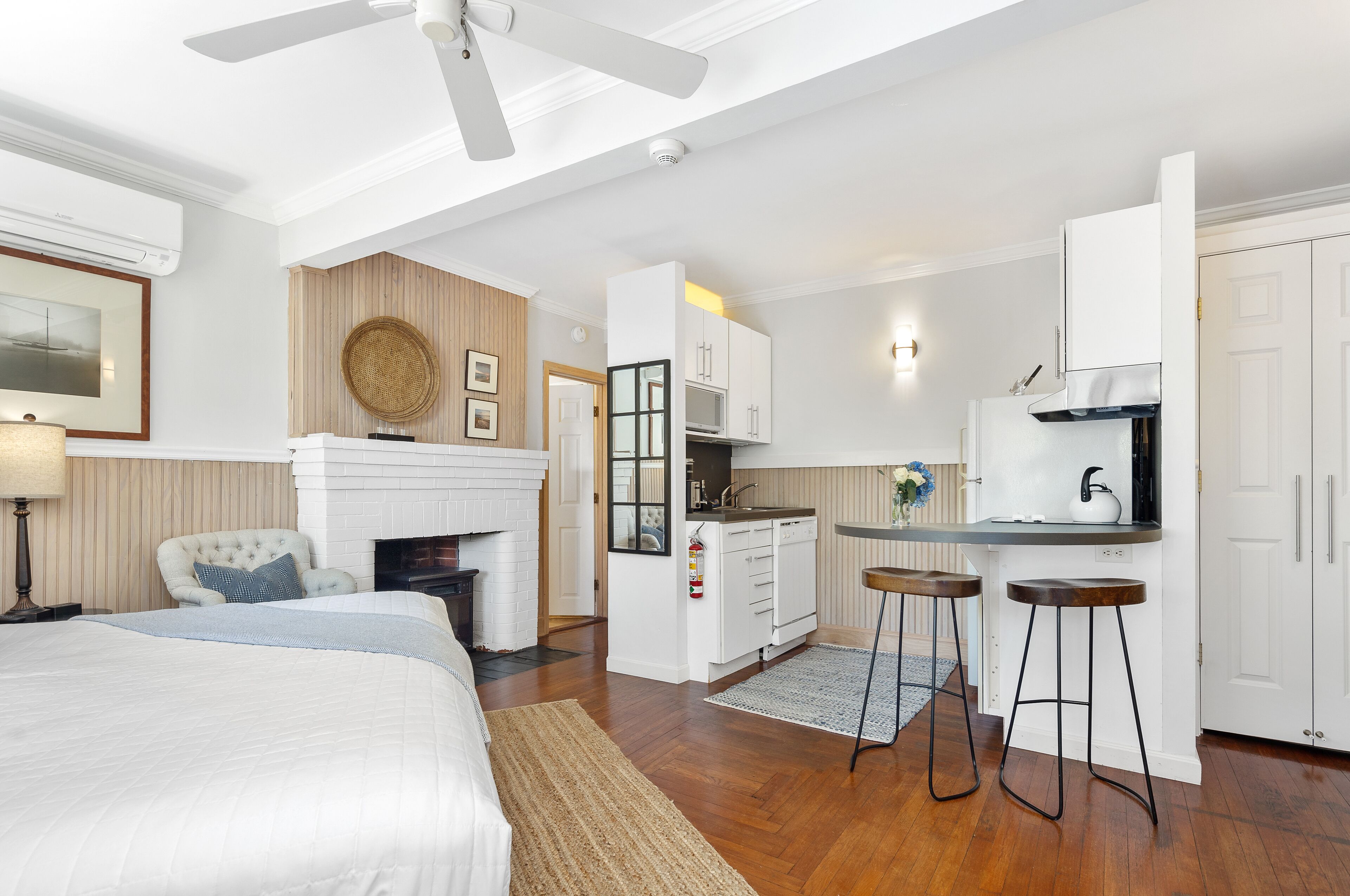 A modern hotel room with a large bed, a sitting area with a chair, a kitchen with white cabinets, and a ceiling fan.