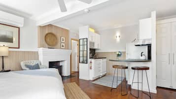 A modern hotel room with a large bed, a sitting area with a chair, a kitchen with white cabinets, and a ceiling fan.