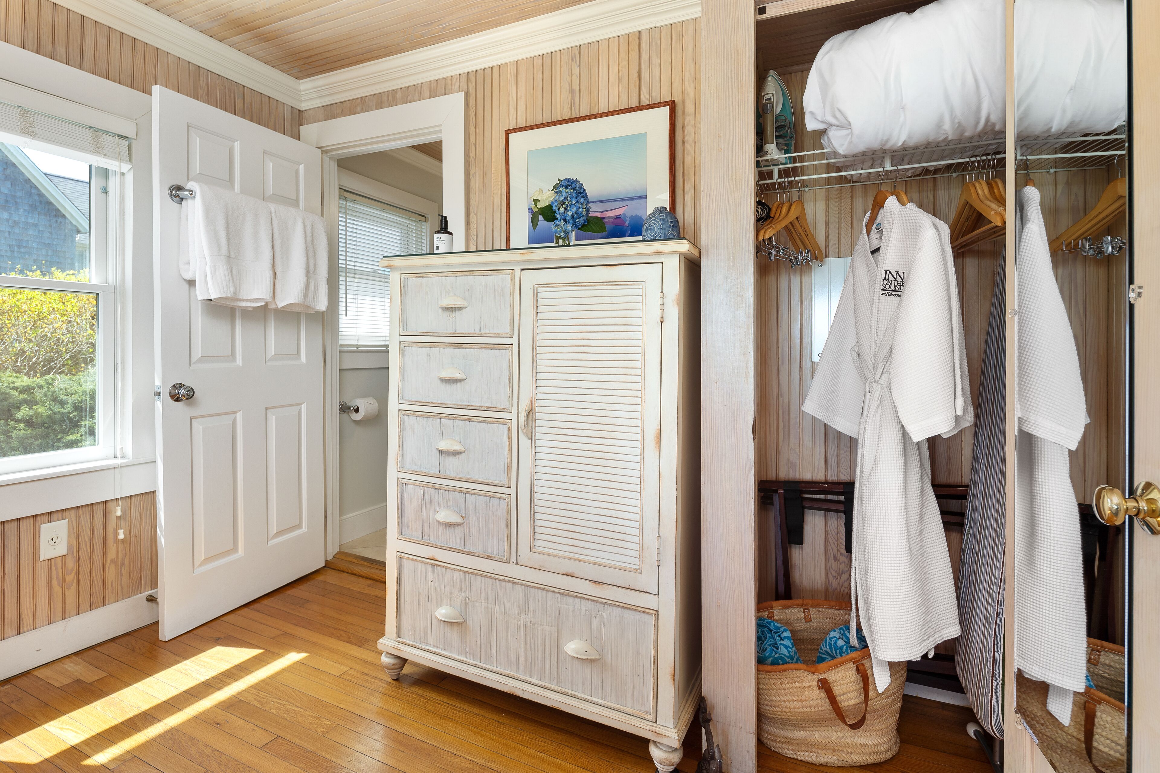 Une pièce avec un plancher en bois, une commode blanche à tiroirs, un placard avec des vêtements suspendus et une fenêtre donnant sur un paysage verdoyant.