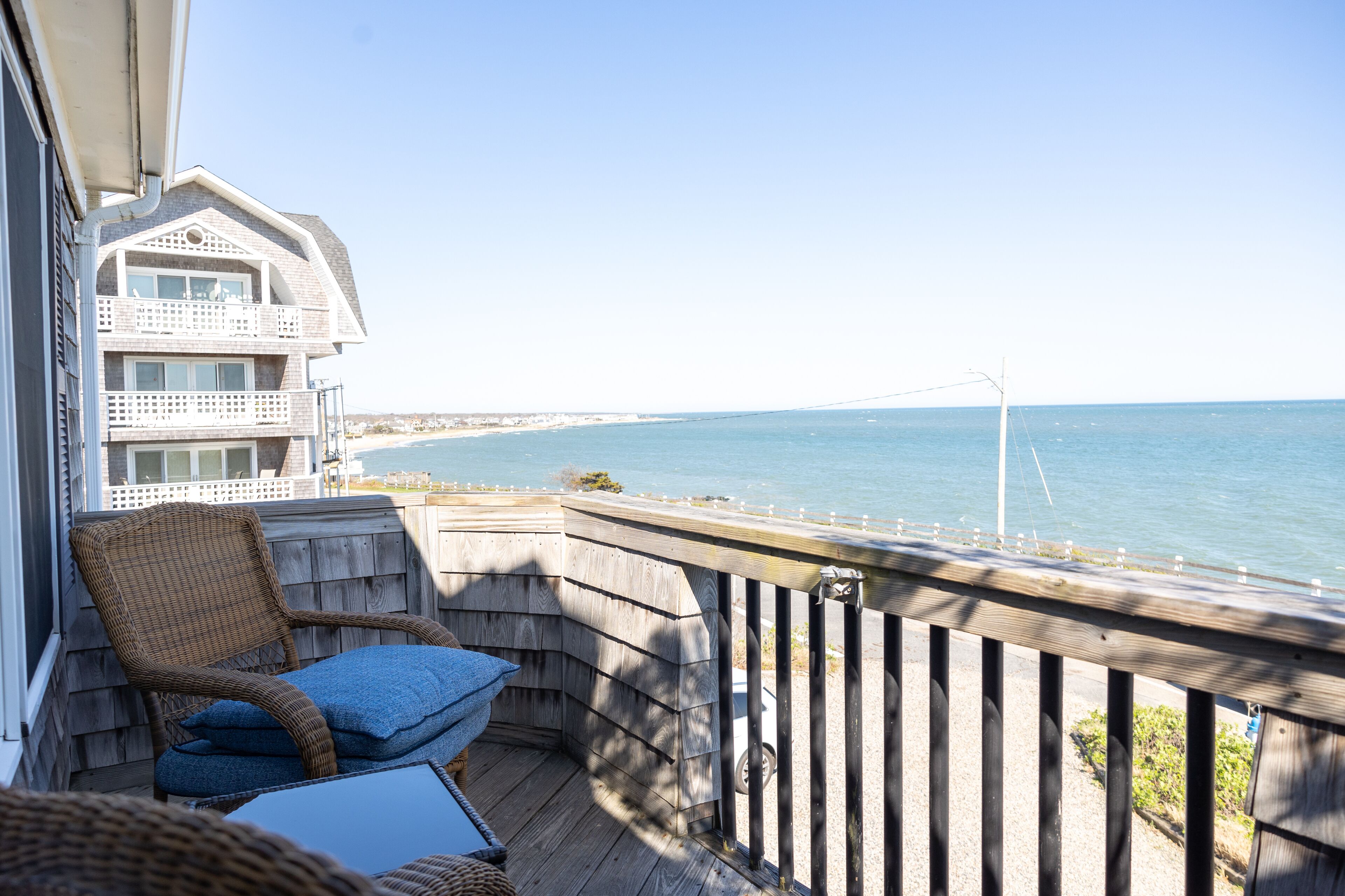A balcony with a wicker chair and cushions, overlooking a beach and a bridge.