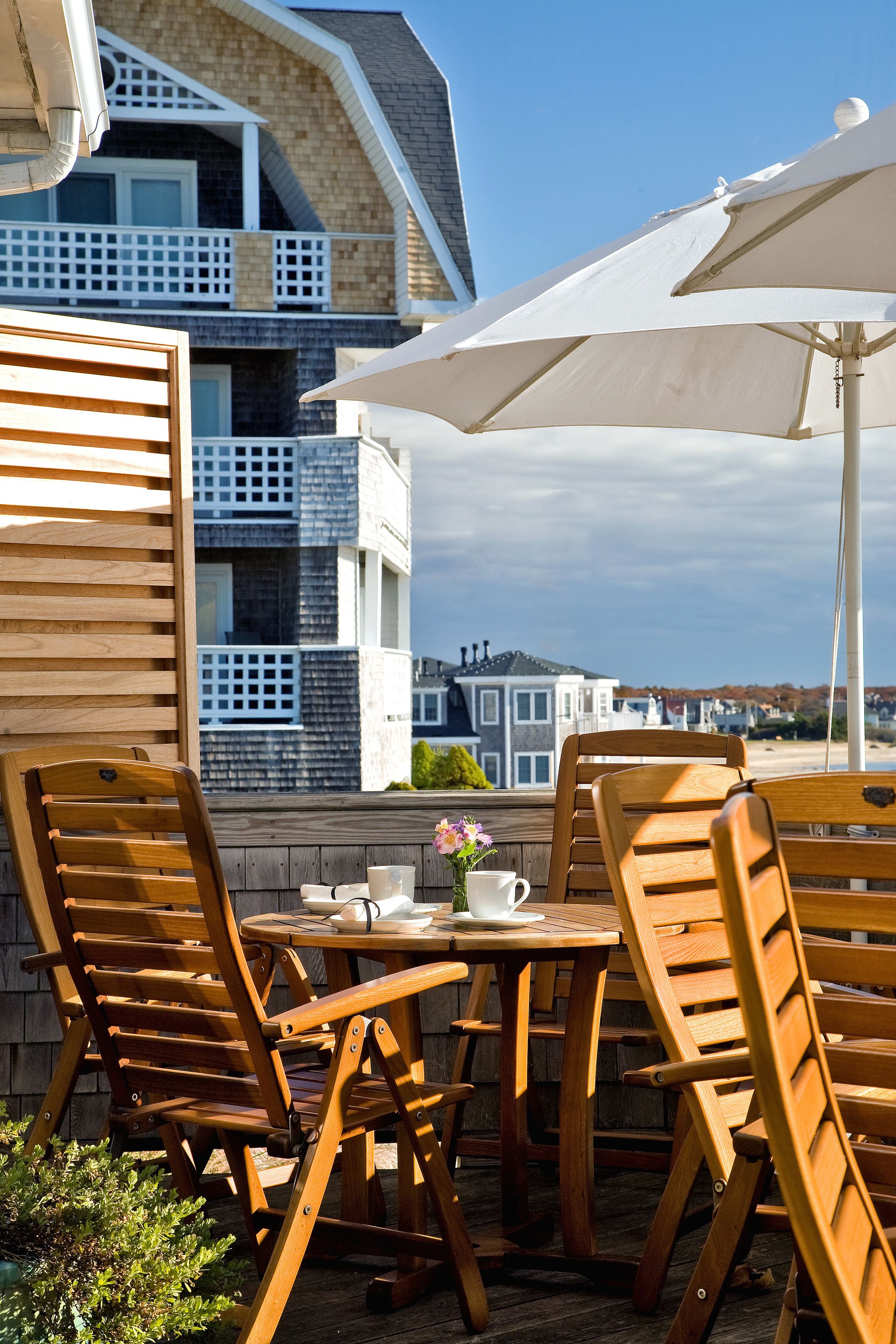 A wooden outdoor table and chairs with a white umbrella, set against a backdrop of a residential building with balconies.