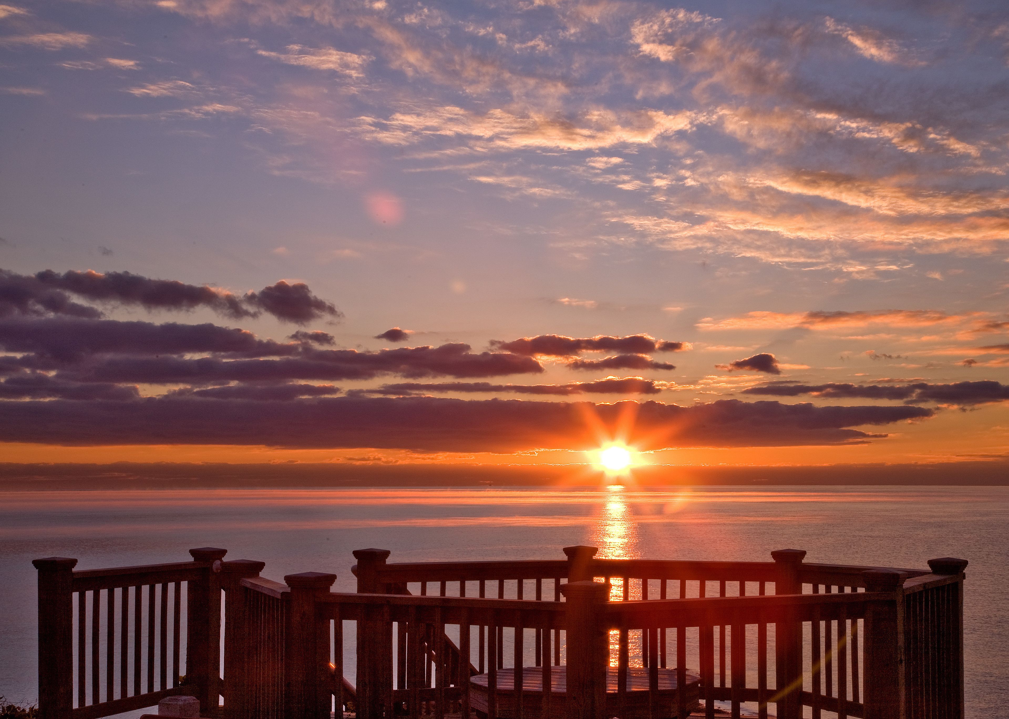 Een zonsondergang boven een wateroppervlak met een houten balustrade op de voorgrond.