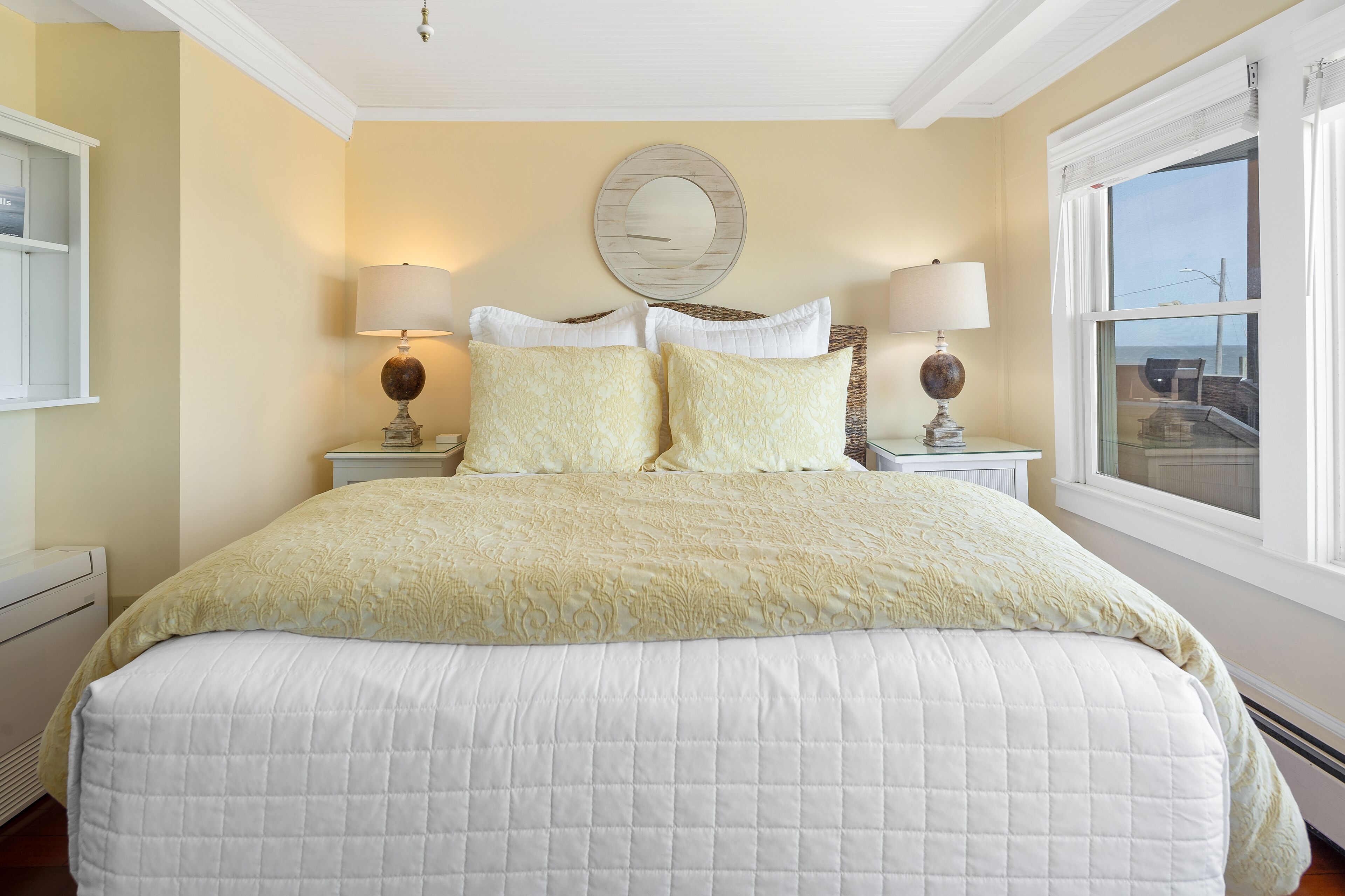 A neatly made bed with a quilted white comforter and two matching pillows, flanked by two bedside tables with lamps, in a room with a large window and a decorative mirror on the wall.