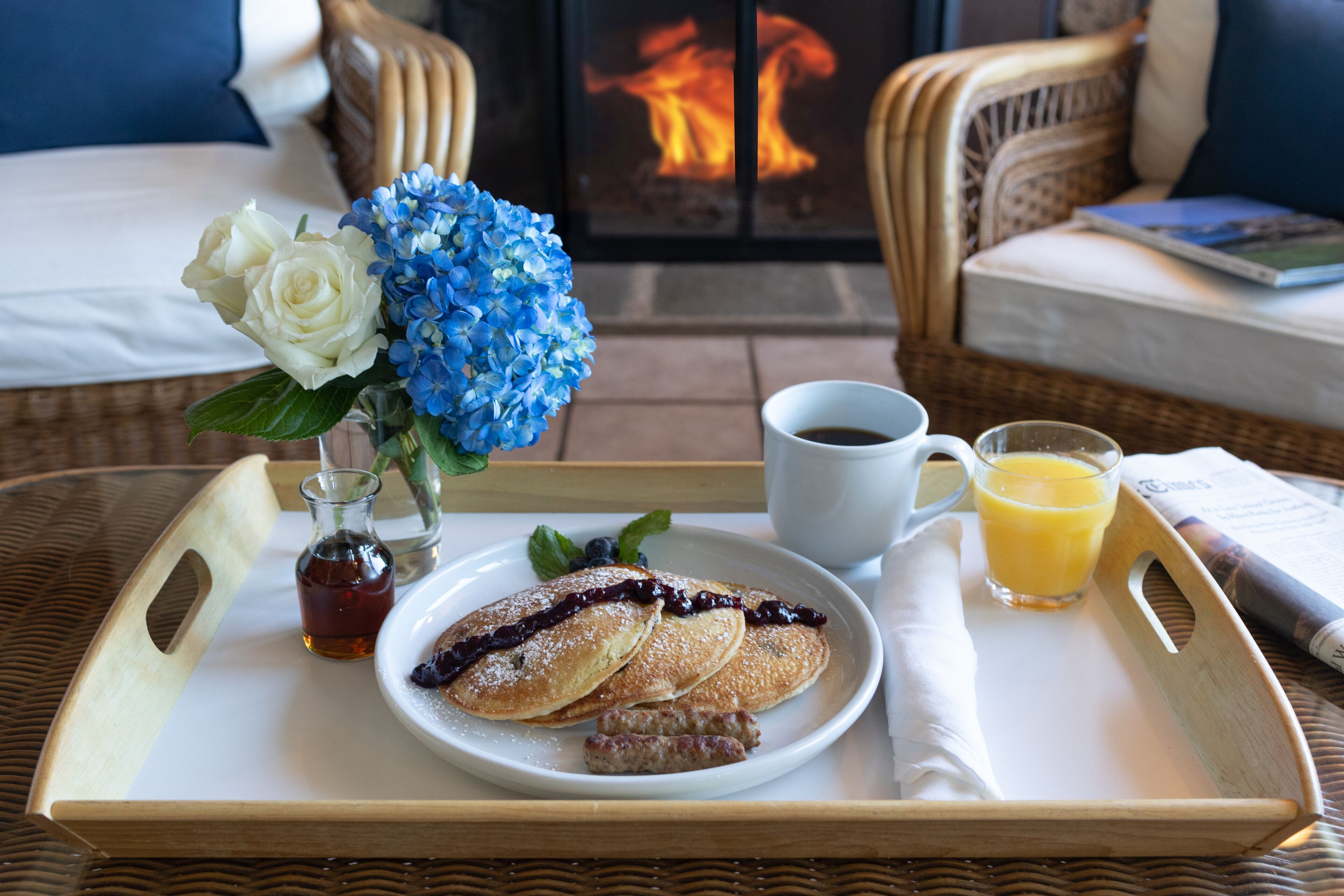 Ein Frühstücksbrett mit Pfannkuchen, Marmelade, Würstchen, einem Glas Orangensaft und einer kleinen Flasche Sirup, platziert auf einem Korbtisch in einem Raum mit Kamin und Korbmöbeln.