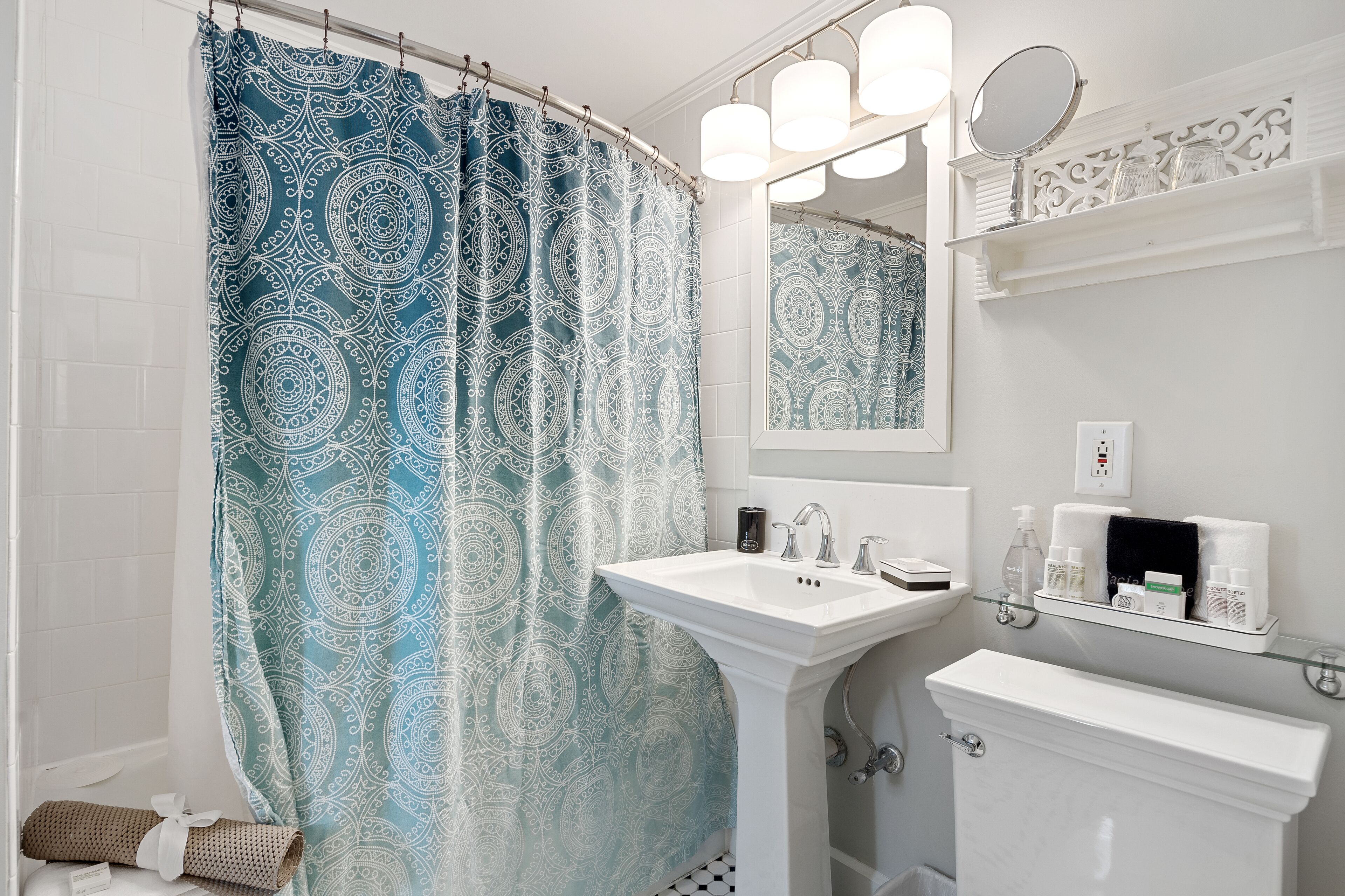 A bathroom with a patterned shower curtain, a pedestal sink, a mirror, and a shelf with toiletries.