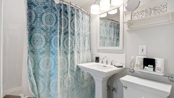 A bathroom with a patterned shower curtain, a pedestal sink, a mirror, and a shelf with toiletries.