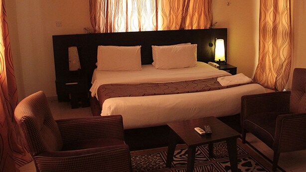 A hotel room with a large bed, two nightstands, a bedside table, two wicker armchairs, and a patterned rug. - Lakeem Suites Adebola (Lagos)