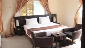 A hotel room with a large bed, two bedside tables, a small table with a chair, and a window with curtains. - Lakeem Suites Adebola (Lagos)