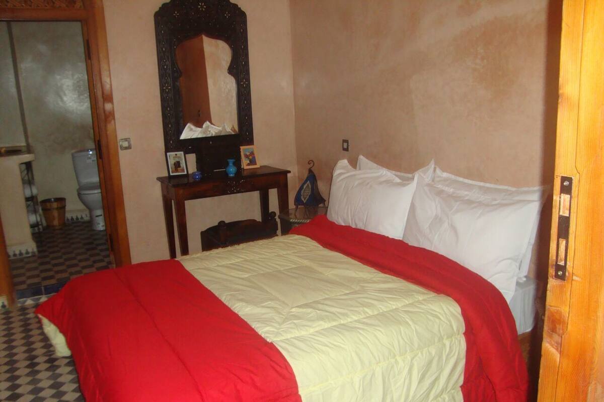 Quarto com cama, mesa de cabeceira de madeira, espelho e porta.