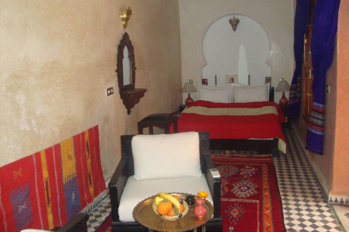 Quarto com uma cama, uma cadeira e um tabuleiro de frutas, além de um tapete estampado.