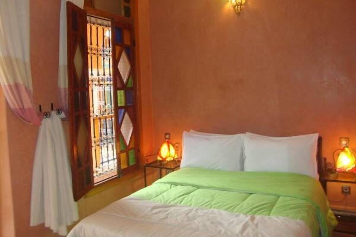 Quarto com cama, duas luminárias de apoio, janela com grade de metal e luminária fixa na parede.