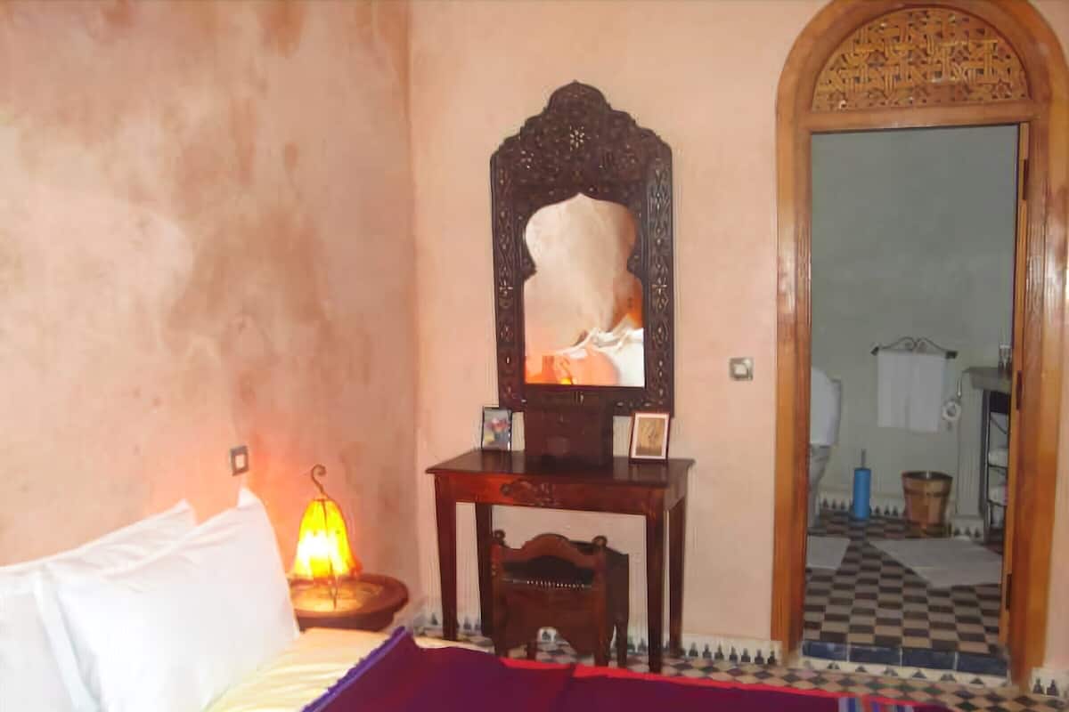 Quarto com cama, abajur, mesa de cabeceira de madeira, espelho e chão quadriculado.