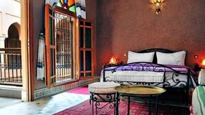 A bedroom with a bed, a nightstand, a small table, a decorative wall lamp, and a view of an outdoor courtyard. - Riad BenSaid (Marrakech)