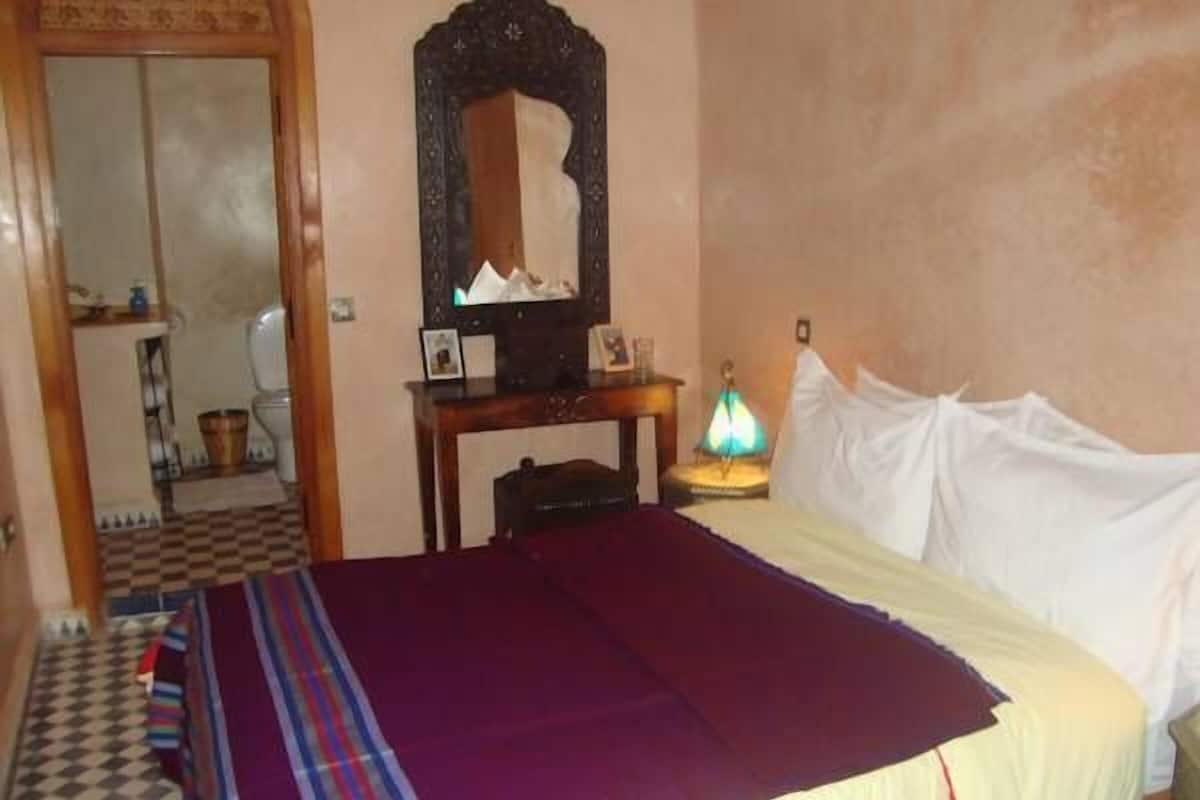Quarto com cama, mesa de cabeceira, luminária, espelho e chão quadriculado.