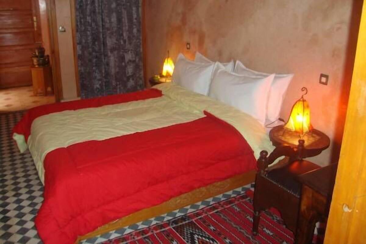 Quarto de hotel com uma cama grande, duas luminárias de cabeceira, piso xadrez e porta de madeira.