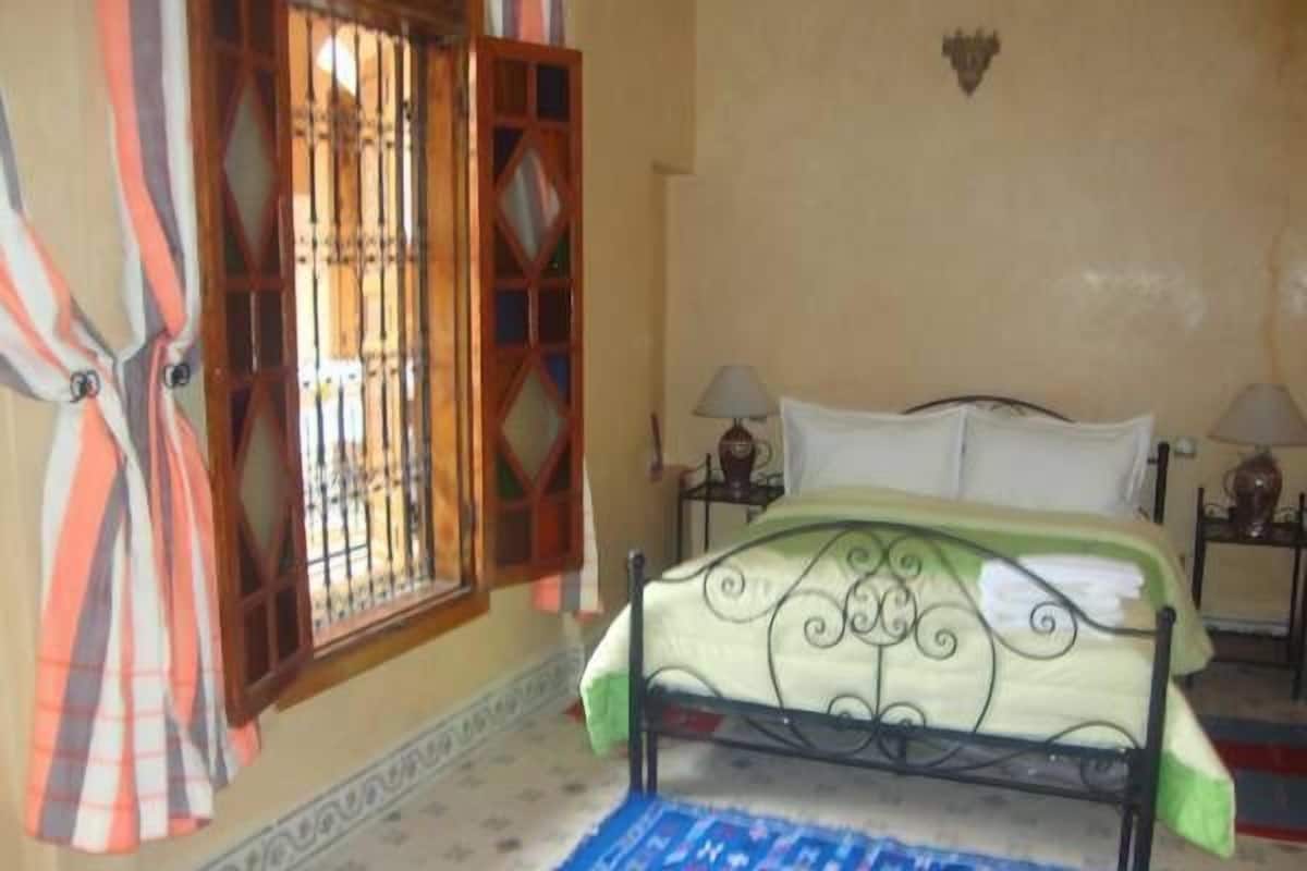 Quarto com cama, duas mesas de cabeceira, janela com grades e cortinas.