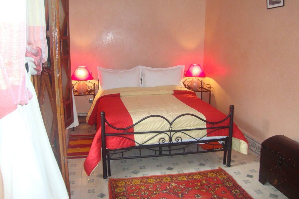 Quarto com cama, duas luminárias de cabeceira, colcha vermelha e branca, tapete estampado e pequeno enfeite de parede.