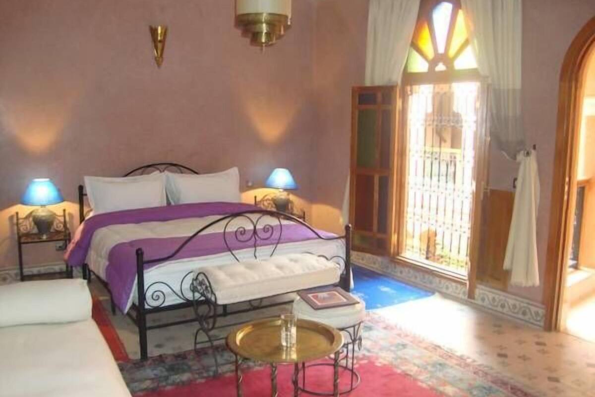 Quarto com cama, duas luminárias de cabeceira, uma mesinha e uma janela com cortinas.