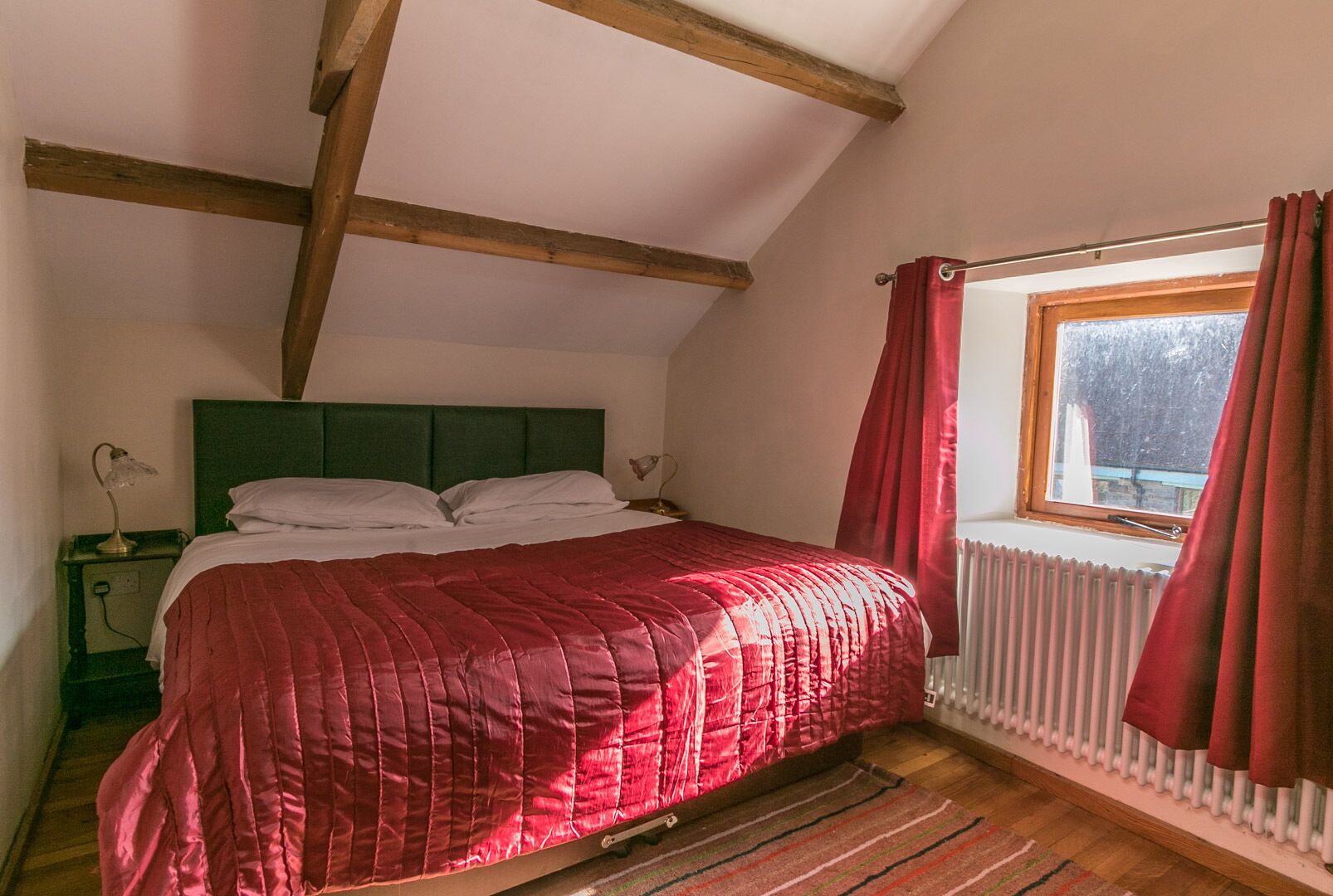 Une chambre à coucher avec un lit, des poutres en bois, une fenêtre avec des rideaux et un radiateur.