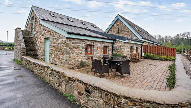 Un bâtiment en pierre avec une terrasse en briques, une clôture en bois et une table entourée de chaises.