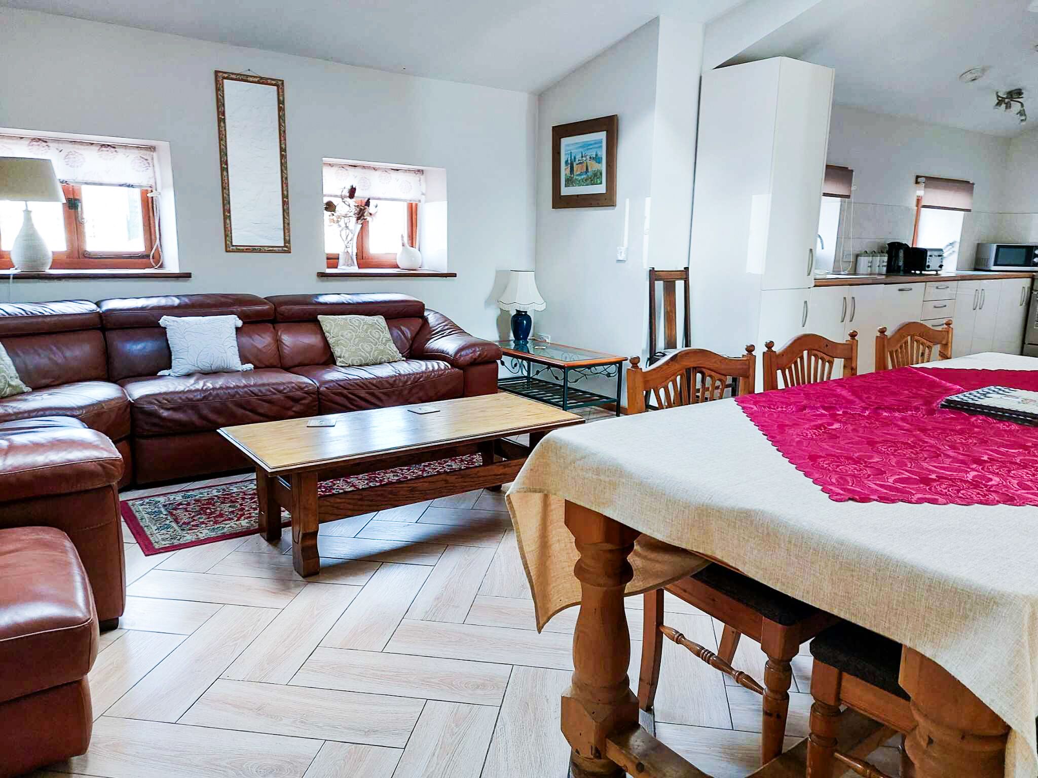 A living room with a brown leather sofa, a wooden coffee table, and a dining table with chairs. There is a kitchen area with white cabinets and a microwave.