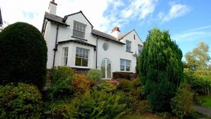 Une maison blanche à deux étages, dotée d’un jardin bien entretenu, comprenant une variété d’arbustes et un grand conifère.