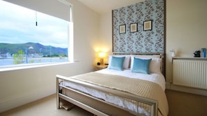 Une chambre à coucher avec un lit, une fenêtre donnant sur un paysage, un mur orné de tableaux encadrés et une table de chevet avec une lampe.